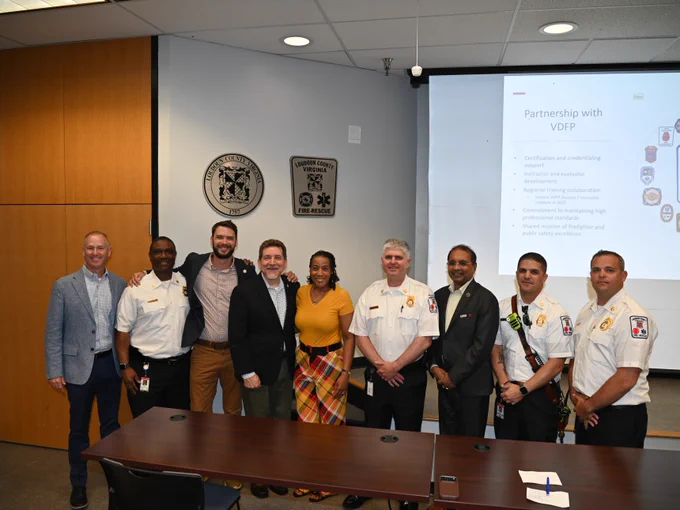 A group of individuals standing in a conference room, some wearing firefighting uniforms with badges. Behind them is a presentation slide titled "Partnership with VDFP" and official seals of government agencies on the wall. They are positioned in front of a wooden table.