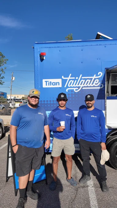 Three people standing in front of a blue "Titan Tailgate" truck, each wearing blue shirts and hats. One person is holding a cup. The background shows a clear sky and part of a building.