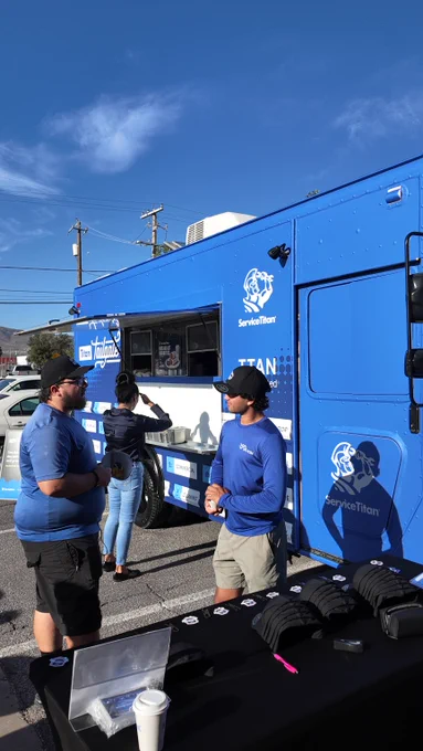 A blue ServiceTitan food truck is parked in a sunny outdoor setting. Two people are standing near the truck, engaged in conversation, while another person is placing an order at the window. A table with promotional items and a coffee cup is in the foreground.