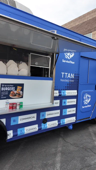 Blue ServiceTitan food truck with logos and text promoting commercial services like pest control and plumbing. Drink cans are on the counter next to a burger poster.
