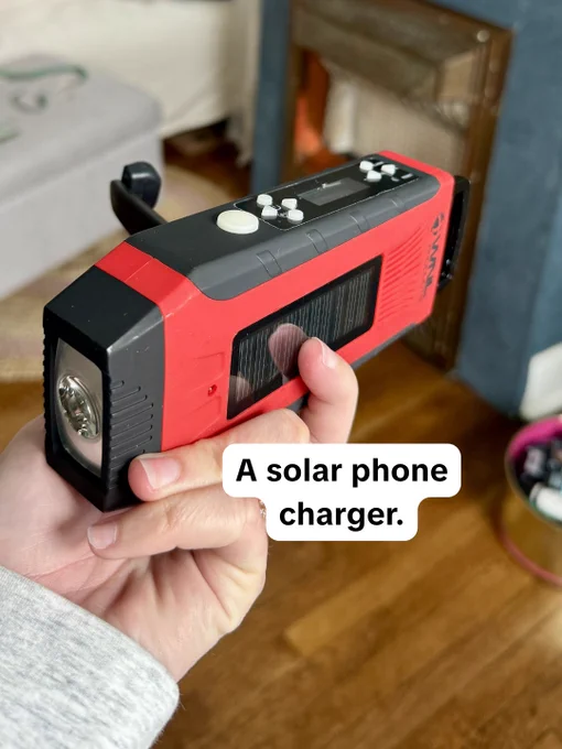 A hand holding a solar-powered phone charger with a red and black design. The device has a crank handle, buttons, and a flashlight. The text "A solar phone charger" is overlaid on the image.