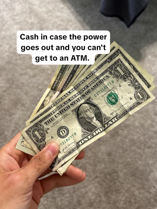 A hand is holding several United States one-dollar bills. A caption above reads, "Cash in case the power goes out and you can't get to an ATM."