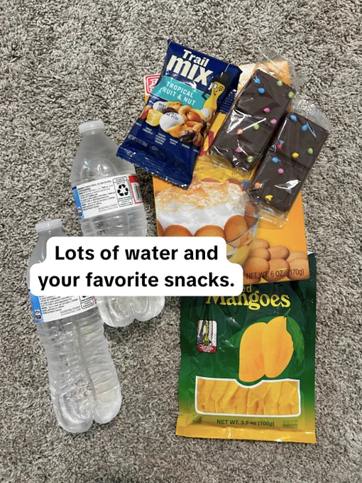 This image shows two bottles of water, a pack of tropical trail mix, a package of mixed fruit bars, and a bag of dried mangoes, arranged on a textured surface. A message reads: "Lots of water and your favorite snacks.