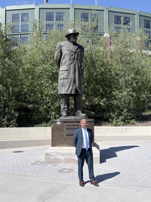 Interim Chancellor Eric Wilcots stands in front of a statue of Vince Lombardi, located at the entrance of Lambeau Field. The statue depicts Lombardi in a long coat and hat. Green trees and part of the stadium are visible in the background.