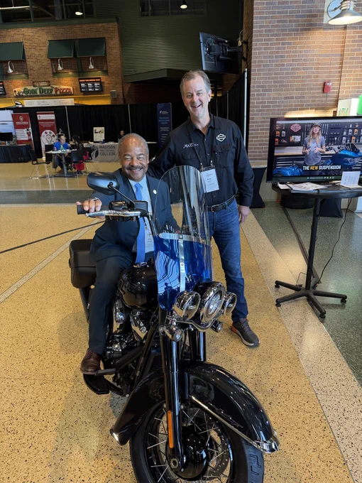 Interim Chancellor Eric Wilcots sitting on a Harley-Davidson motorcycle inside a spacious, well-lit room. Harley CEO Artie Starrs stands beside them, both smiling. In the background, there's a booth with Harley-Davidson branding. 