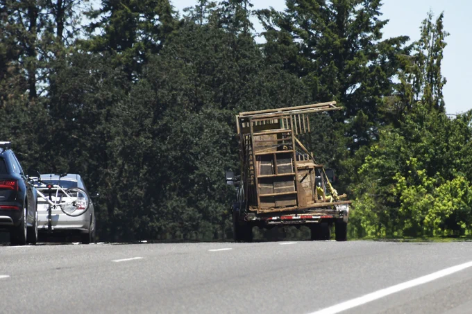 A truck overloaded with large wooden furniture drives on a road surrounded by tall trees. Two cars are parked on the side of the road, with one carrying bicycles on a rear rack. The sky is clear and sunny.