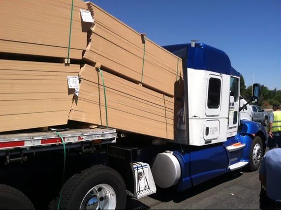 A blue and white semi-truck with large stacks of wooden boards on the trailer that have slid off and into the driver's cabin. An individual in a reflective vest stands nearby, observing the scene.