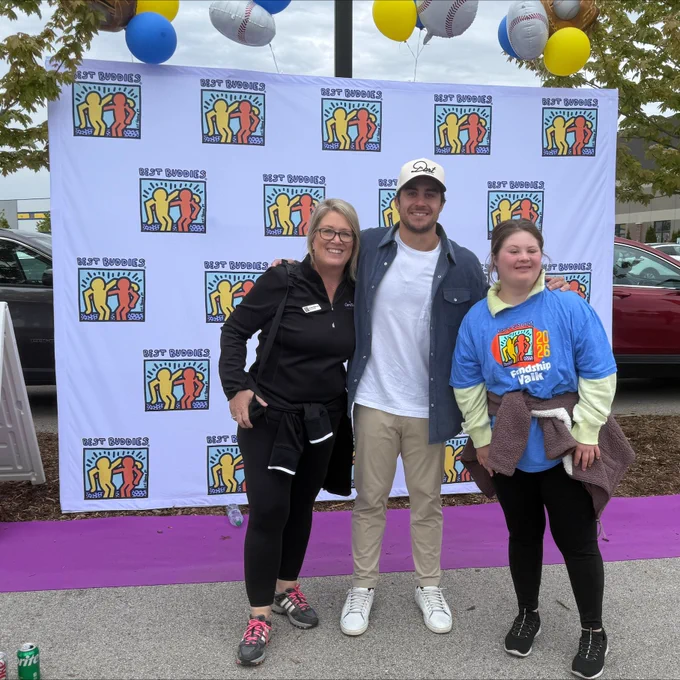 A group of three people standing in front of a backdrop, decorated with balloons. There's a purple carpet on the ground and a car and trees in the background.