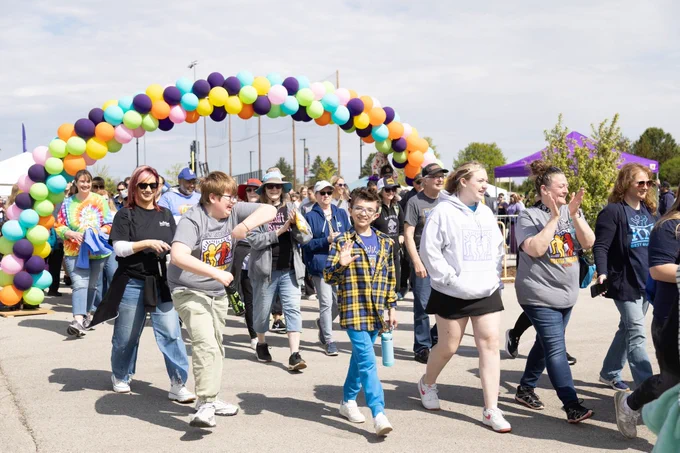 A group of people are participating in a walk event, moving under a colorful balloon arch. Participants are wearing casual clothing, some in event t-shirts. The atmosphere appears lively and cheerful.