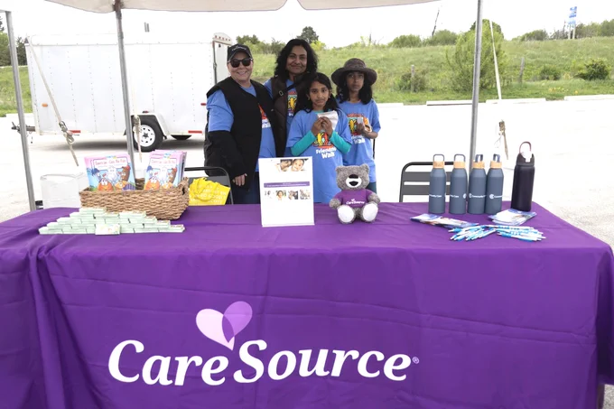 A group of people stands behind a table draped with a purple cloth displaying the "CareSource" logo. The table holds brochures, a stuffed bear, water bottles, and other promotional items. There is a canopy above, and a trailer is visible in the background.