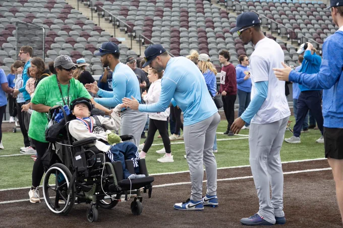 A person in a wheelchair on a baseball field is interacting with athletes, who are wearing athletic gear.