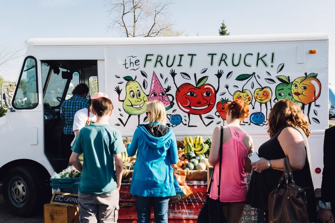 Four people are shopping at a white food truck that reads “The Fruit Truck” with illustrations of fruits and vegetables. 