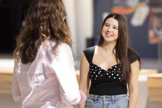 Two people are conversing indoors. One wears a black and white polka dot top and jeans, while the other is dressed in a light shirt with long hair. Background includes blurred art pieces and a large pillar.