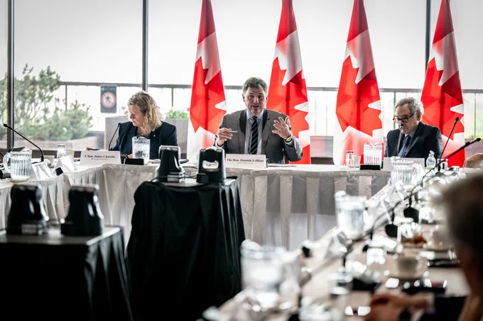 Le ministre LeBlanc prend la parole lors d’une réunion du Comité consultatif sur les relations économiques entre le Canada et les États-Unis, aux côtés de la négociatrice en chef pour les États-Unis, Janice Charette, et d’autres participants, avec des drapeaux du Canada en arrière-plan.