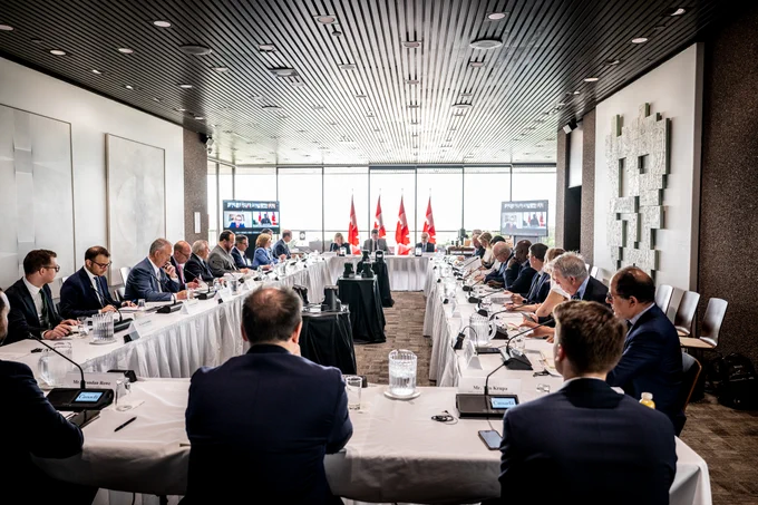 Les membres du Comité consultatif sur les relations économiques entre le Canada et les États-Unis participent à une réunion autour d’une table de conférence afin de discuter des priorités commerciales et économiques entre le Canada et les États-Unis, avec des drapeaux du Canada à l’avant de la salle.