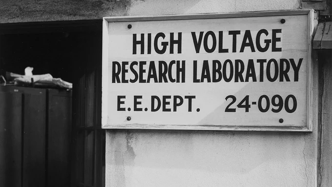Black-and-white photo of a sign labeled 'High Voltage Research Laboratory E.E. Dept. 24-090' mounted next to a doorway.
 