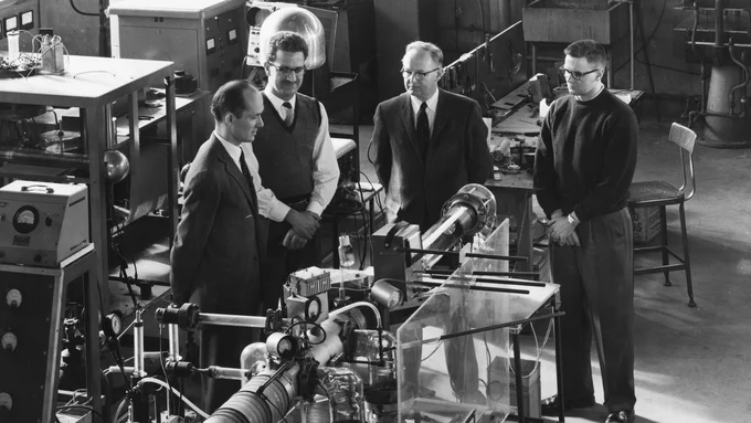 Four men, John G. Trump is second from the left, are in a laboratory examining experimental equipment, likely part of a scientific research team. The room is filled with various scientific instruments and machinery.
