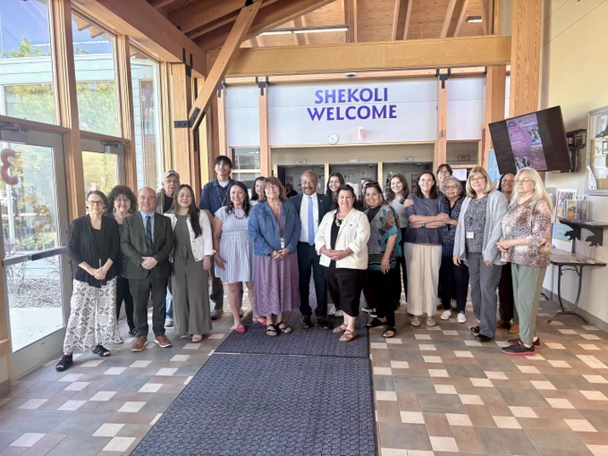 A group of people including interim Chancellor Wilcots standing in a lobby area with a "SHEKOLI WELCOME" sign above. They are posed together on a tiled floor, and there is natural light coming in through large windows.