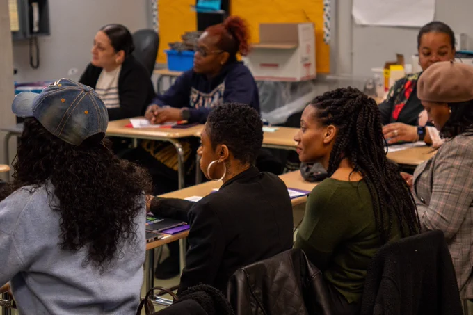 A diverse group of adults attentively listening in a classroom setting with desks and educational materials.