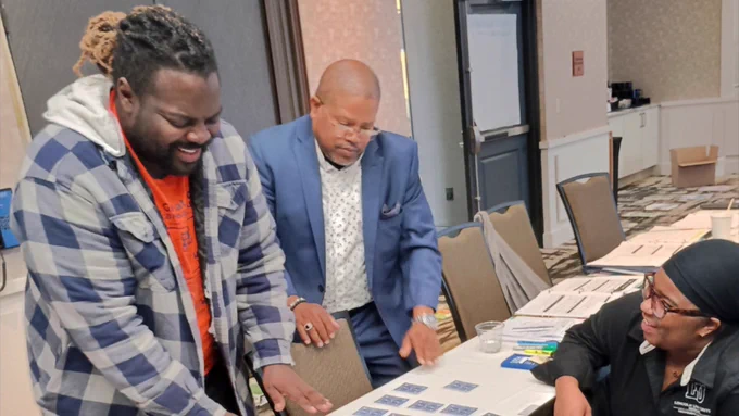 An image from the Lincoln University 4-H staff retreat showing three educators participating in a hands-on card-matching activity at a long conference table. In the foreground, a man in a blue and white plaid jacket smiles as he reaches toward face-down cards on the table, while a man in a blue suit stands next to him watching closely. On the right, a woman in a black Lincoln University shirt looks up and smiles.
