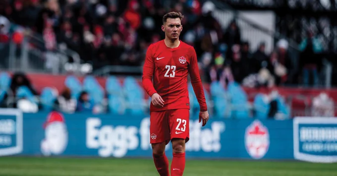 A soccer player wearing a red Canada national team jersey with the number 23 walks on the field. In the background, there are empty blue seats and blurred spectators. Advertisements, including a visible Canadian soccer logo, are displayed around the field. Image credit: Canada Soccer.