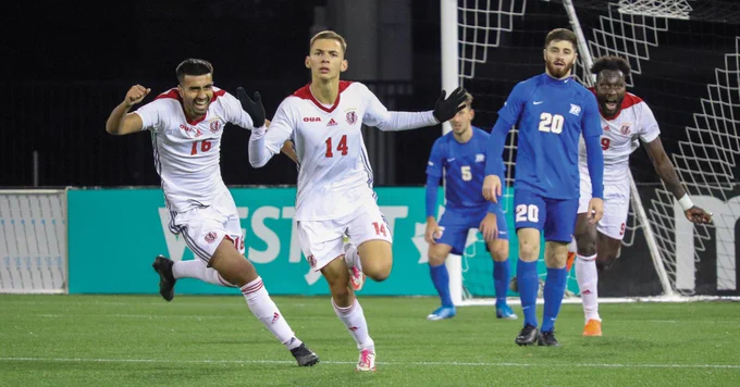 Soccer players on a field are celebrating a goal. Two players in white jerseys appear happy and are running, while two players in blue jerseys look on. A goal net is in the background.