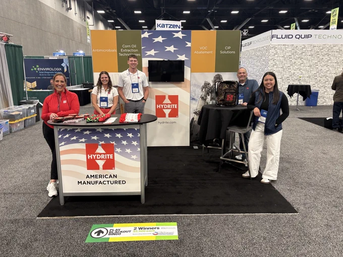 Four people stand and sit around a Hydrite booth at a trade show, featuring American flag branding and product displays.