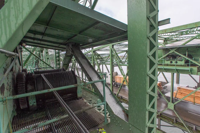 The image shows a close-up of the intricate green metal framework of the Interstate Bridge. Large gears and cables that power the bridge's lift span are visible among the bridge's structural and mechanical components. In the background, cars and a roadway are visible, with the Columbia River under the bridge. 