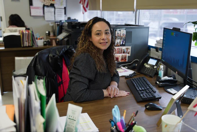 Woman with curly hair sitting at an office desk with a computer, phone, and various papers in a bright workspace.