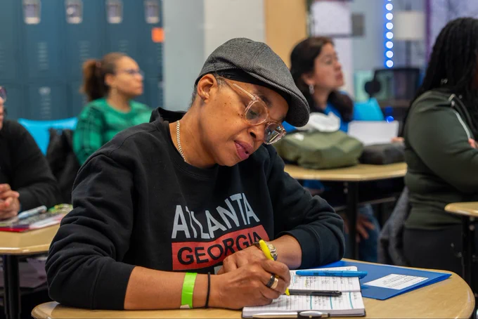 Woman wearing an Atlanta Georgia sweatshirt and a cap, focused on writing notes in a notebook at a classroom desk.