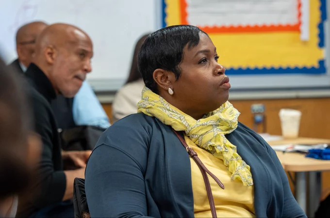 A woman wearing a yellow scarf and blue cardigan attentively listens during a classroom discussion with other adults in the background.