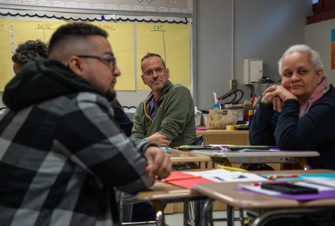 A group of adults engaged in a classroom discussion, seated around desks with notebooks and papers.