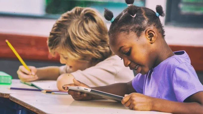 Menina negra usa tablet sorrindo, sentada ao lado de menino loiro que escreve em caderno com lápis amarelo em sala de aula.