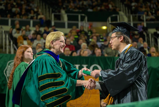 University president shaking the hand of a graduate and handing over a diploma.