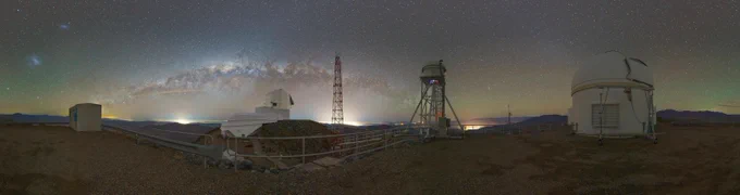 Panoramic night view of a mountaintop observatory site. From left to right we see: a metal storage container under the two ghostly smudges of the Large and Small Magellanic Clouds, a boot-shaped observatory building next to a red antenna beneath the Milky Way arching over a star-filled sky, a tall spindly tower topped with a small dome, and a larger traditional cylindrical telescope building.
