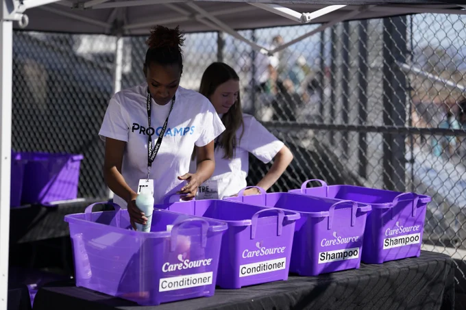 Two individuals are organizing purple bins labeled "CareSource Conditioner" and "CareSource Shampoo" under a tent. They are wearing white "ProCamps" shirts.
