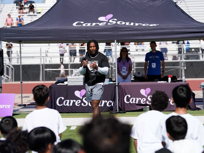 A person stands on a sports field in front of a group of people. They are speaking into a microphone under a canopy bearing the logo "CareSource." The audience, dressed in white shirts, is gathered closely. Two other individuals are seated at a table to the side under the canopy.