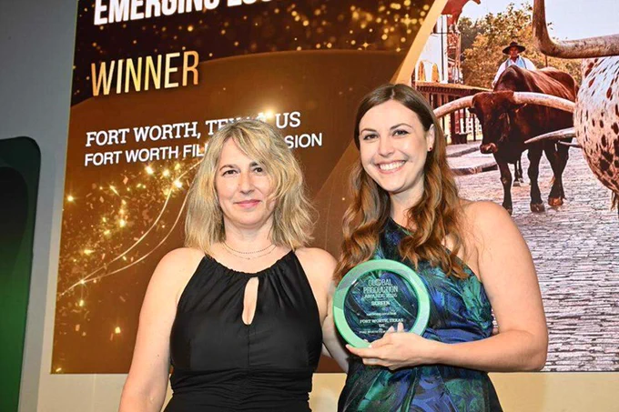 Two people standing and smiling at an award event. One holds a circular award. A presentation screen in the background reads "Emerging Destinations Winner, Fort Worth, Texas, US, Fort Worth Film Commission." A cowboy and longhorn cattle are also visible in the projected image.