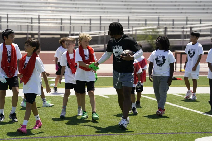A group of young players on a football field, wearing athletic gear. One person is holding a football and interacting with the group. Some players have red practice vests, and bleachers are visible in the background.