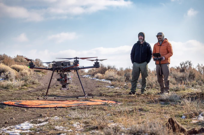 Two men stand in a dry, grassy field operating a large professional drone on an orange landing pad under a partly cloudy sky.