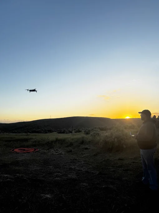 Person operating a drone flying over a grassy field at sunset with hills in the background.