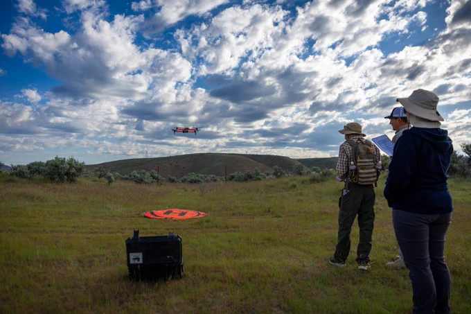 Three people operating a drone flying over a grassy field under a partly cloudy sky.
