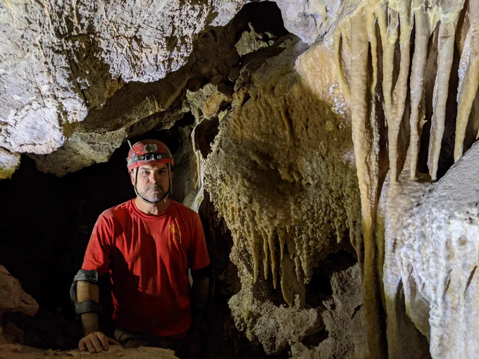 Person wearing a red shirt and headlamp exploring a dimly lit cave with prominent stalactites and rugged rock formations.