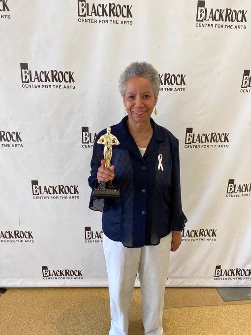 Person holding a golden trophy standing in front of a Black Rock Center for the Arts backdrop, wearing a navy shirt and white pants.