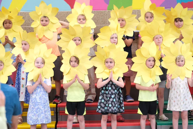Group of children standing on colorful steps wearing large yellow sunflower headpieces, posing with hands together in front of them.