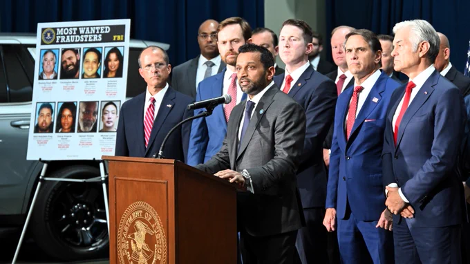 FBI Director Patel speaks at a podium with a Most Wanted Fraudsters FBI board displayed in the background.