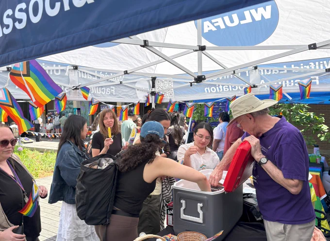 Attendees gather under WLUFA and WLUSA tents decorated with Pride flags while volunteers hand out popsicles.