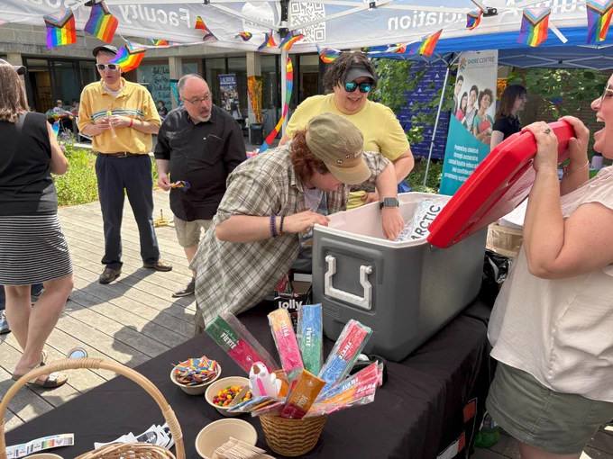 A lively Pride and Popsicles table with attendees choosing treats from a cooler, surrounded by Pride flags and colourful popsicles.