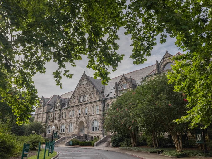 A large, historic stone building, identified as part of Tulane University, is framed by lush green trees. The building features arched windows and intricate architectural details. A pathway leads up to the entrance, and the scene is calm and inviting.