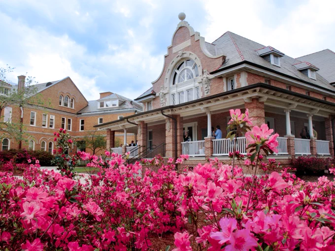 A historic brick building with a large arched window, surrounded by vibrant pink azalea flowers in the foreground. The structure features a spacious porch with white railings. The sky above is partly cloudy, adding to the scenic view.